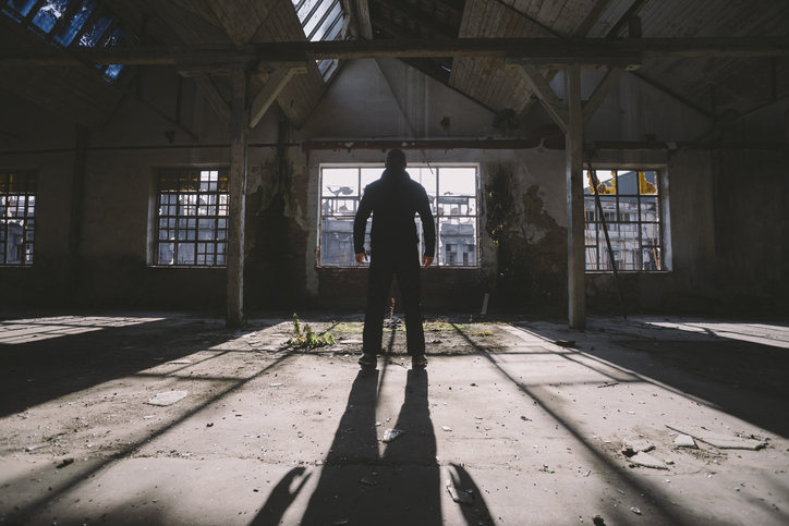 Adult man standing inside some large, dark, spooky,abandoned building illuminated with sunlight through window