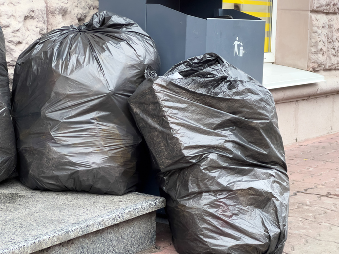 Black garbage bags are piled on the sidewalk beside a public trash can in a busy urban setting, capturing daily waste disposal